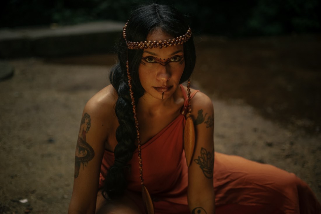 Marcele sentada no chão de terra, envolta em tecido terracota, com trança longa e tiara de contas.