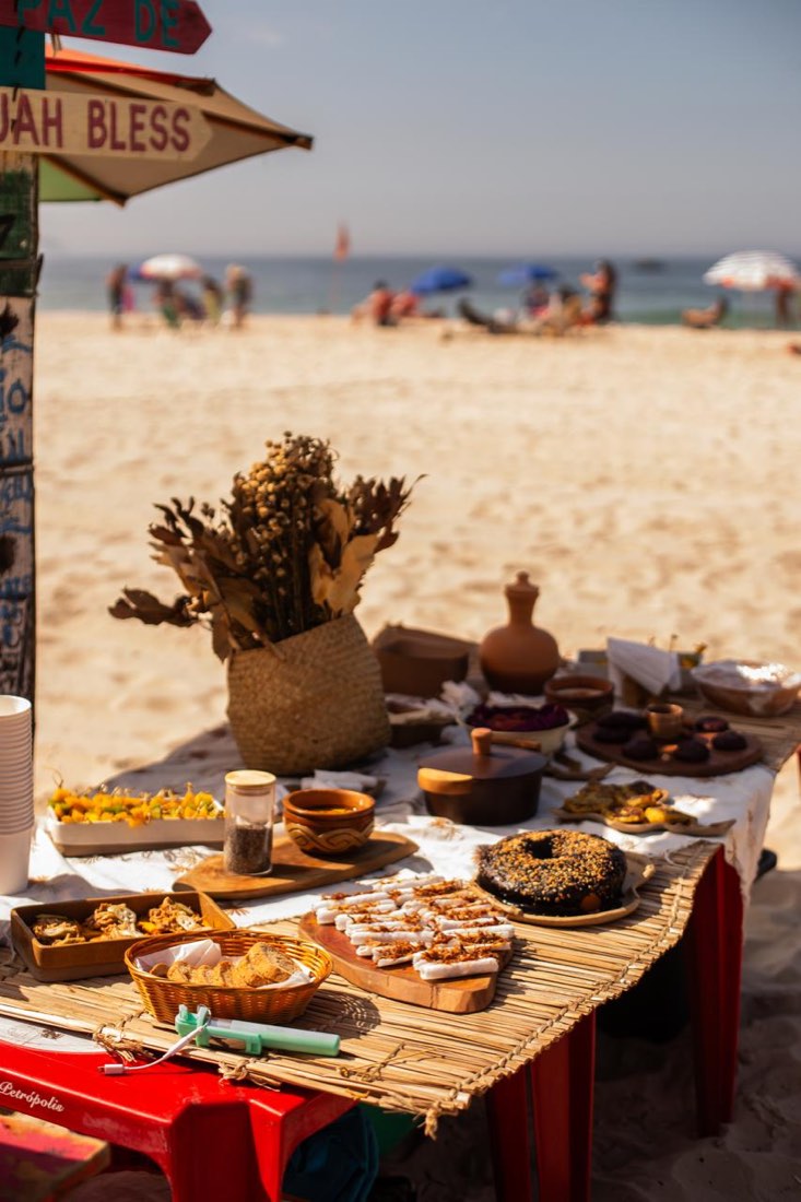 Mesa de café da manhã afro-brasileiro montada na areia da praia do Leme, com pães, bolo, cerâmica e flores secas, sob a placa Jah Bless.